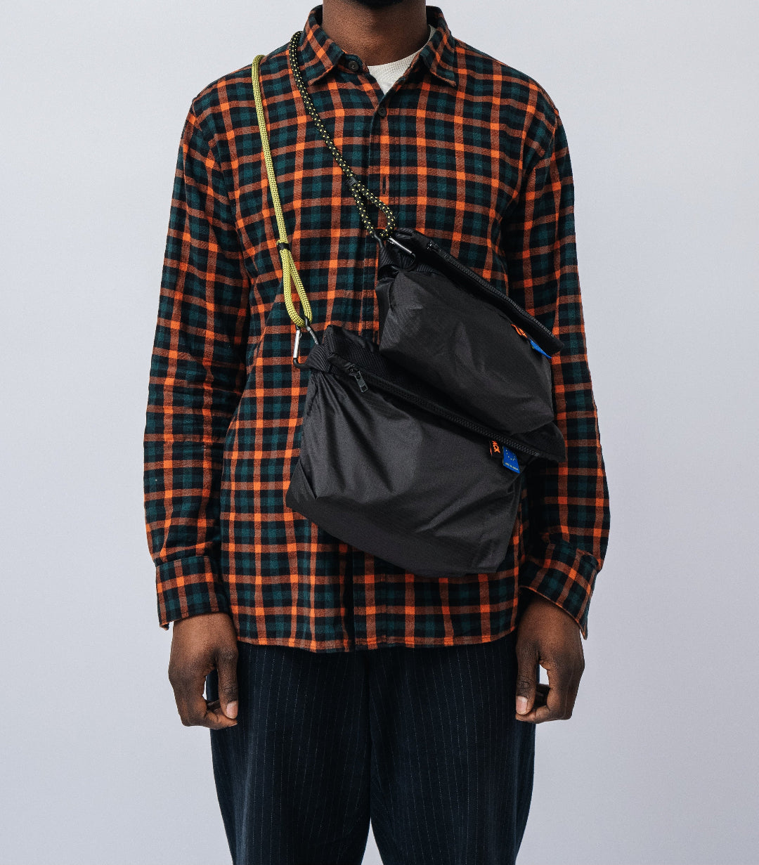 Man wearing a plaid shirt with a black crossbody bag on a plain background